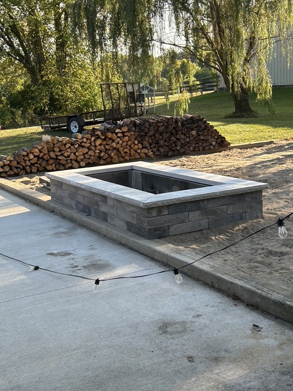 Square gray block fire pit beside a concrete patio with string lights