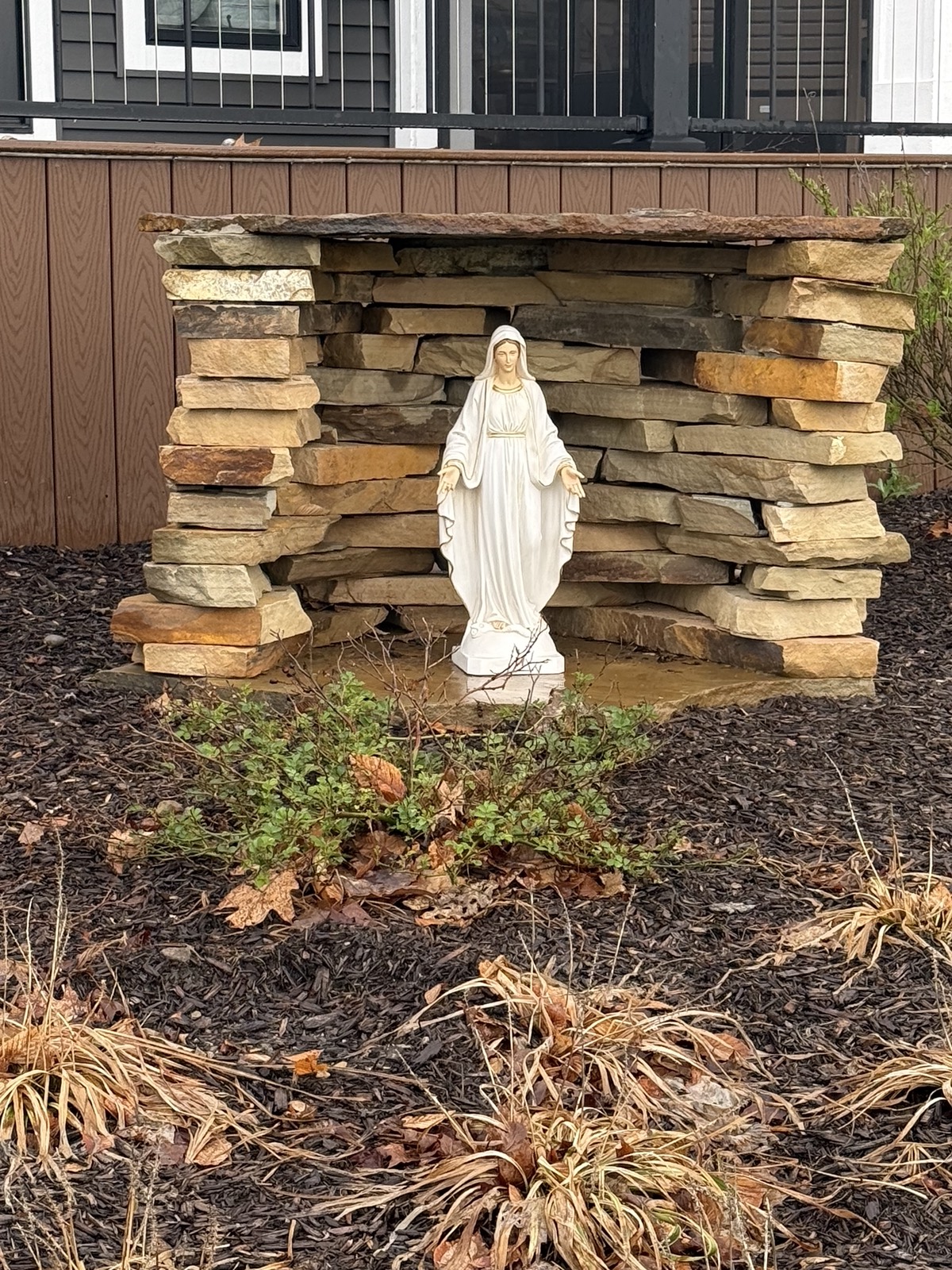 Finished stacked-stone grotto with a white Mary statue, set in a mulched garden bed